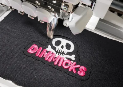 Broderie textile personnalisée en Ariège 6 Logo The Dimmicks rose et blanc en cours de broderie sur textile noir